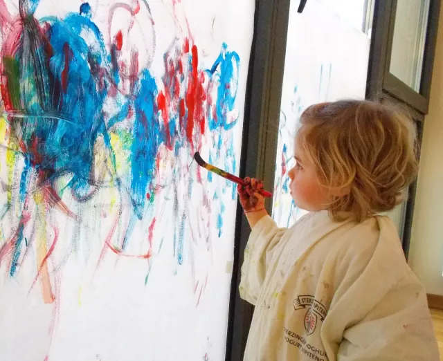 bambina impugna un pennello colorando un grande foglio verticale dipinto con molti colori 