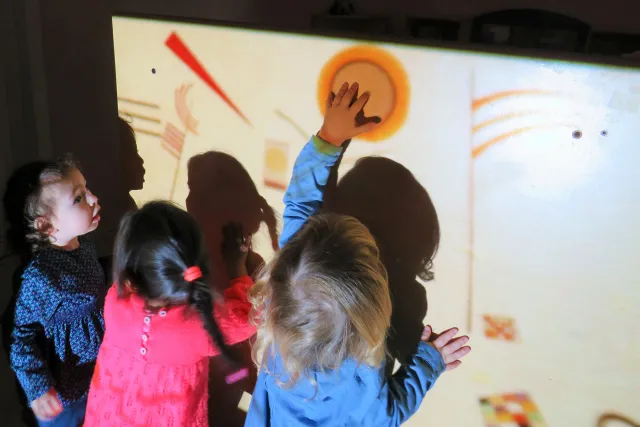 tre bambine osservano una parete con proiezioni luminose di un dipinto. Una tocca con la mano l'immagine del sole