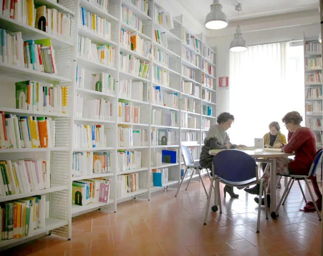 Interno della Biblioteca con libreria a parete ricolma di libri, un'ampia finestra luminosa e tre utenti in consultazione 