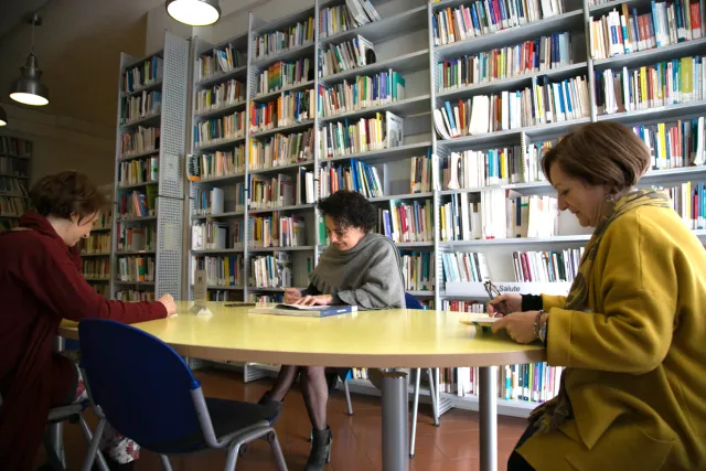 Interno della Biblioteca con libreria a parete ricolma di libri e tre utenti in consultazione attorno a un tavolo