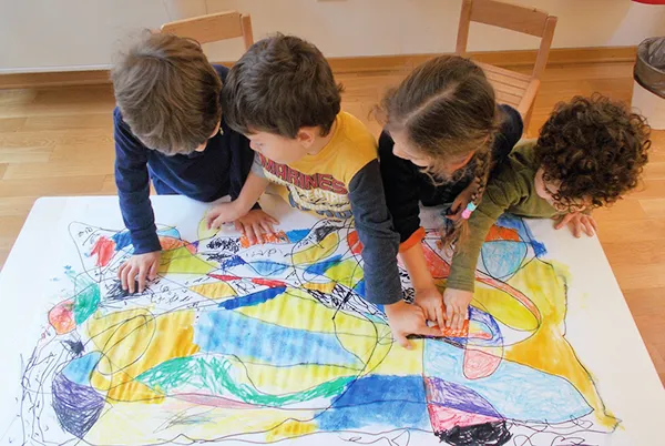 Quattro bambini inginocchiati sui margini di un grande foglio disegnato indicano i diversi colori
