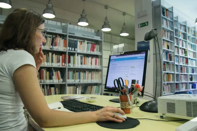 Una donna seduta a un tavolo di fronte a un computer portatile, all'interno di una biblioteca con librerie colme di libri