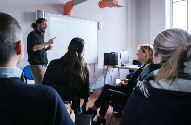 Un docente in piedi, affiancato da una lavagna luminosa, conduce una lezione rivolto a un gruppo di ascoltatori seduti