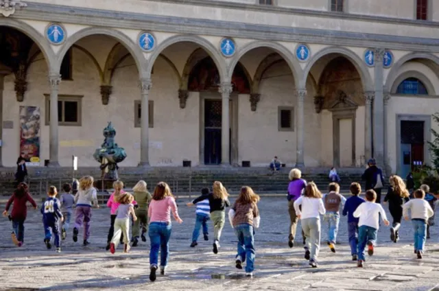 Numerosi bambini, visti di spalle, corrono in piazza verso la facciata con loggiato dell'Istituto