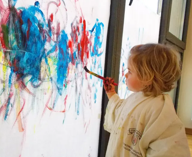 bambina impugna un pennello colorando un grande foglio verticale dipinto con molti colori