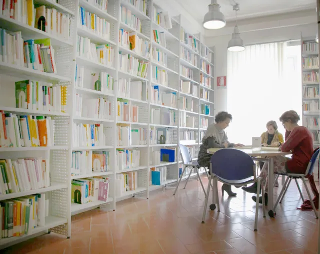 Interno della Biblioteca conuna una libreria a parete ricolma di libri, un'ampia finestra luminosa e tre utenti in consultazione attorno a un tavolo