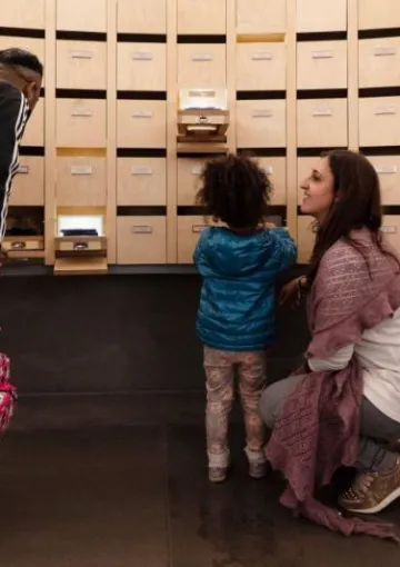 un uomo, una bambina e una donna osservano una sala espositiva del Museo degli Innocenti con cassettini estraibili