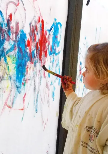 bambina impugna un pennello colorando un grande foglio verticale dipinto con molti colori 