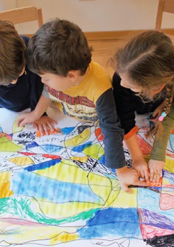 Quattro bambini inginocchiati sui margini di un grande foglio disegnato indicano i diversi colori